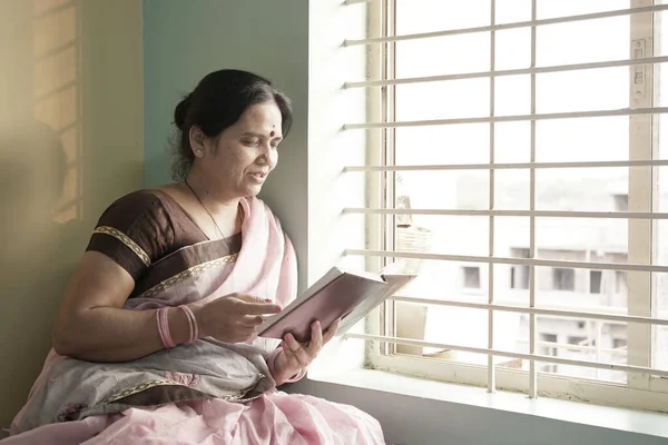Woman staying home for safety during coronaviru pandemic, reading book near window.