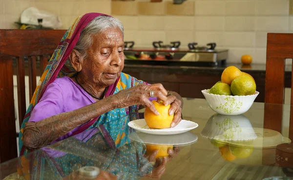 Old Indian woman eating healthy Orange on breakfast table.