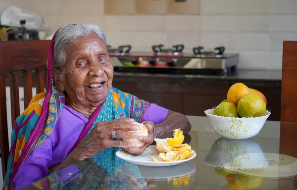 Old Indian woman eating healthy Orange on breakfast table.