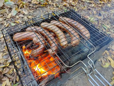 Büyük et sosisleri ızgarada kızartılıyor, yakın plan ateşte. Yaprakların ızgarasının altında bulanık bir arka planda