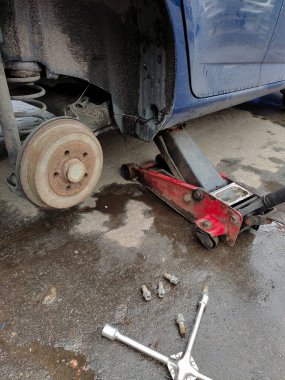 Arka tekeri sökülmüş bir araba kriko ile kaldırılır. Ceviz ve anahtar ıslak asfalta saçılmış. Arabanın bir kısmı görünüyor.