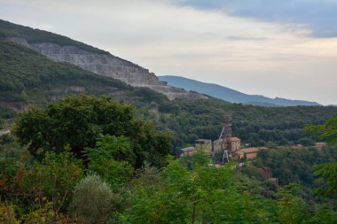 On the mountain slopes there are traces of mining of minerals. In the foreground is an old quarry with buildings in the middle of a green forest