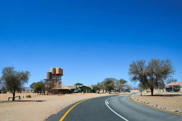 青い空の背景に砂漠の集落の観点からアスファルトの道路 小さな家 木の近くの水の塔 — ストック写真