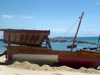 Çatılı eski boş bir balıkçı teknesi kumlu bir sahilde yan yatıyordu. Arkasında mavi gökyüzü ve deniz var.