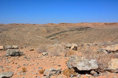 Çöl tepelerine farklı boyutlarda taşlar serpiştirilmiş yamaçlara ve ufukta mavi gökyüzüne.