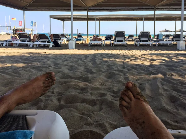 The feet of a man and a woman on the background of a sea beach with lounge chairs of vacationers, the sea in the distance and pavilions from the sun