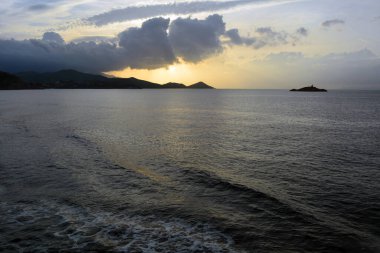 Bulutların arkasında deniz batımı. Uzakta gölgeli dağlar ve zar zor görülebilen küçük bir deniz feneri olan bir ada var.