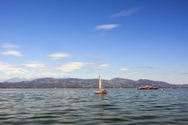 Yatlar ve küçük bir gemi uzak dağların arka planına ve mavi gökyüzüne karşı denizi aşıyor. Yaz deniz manzarası