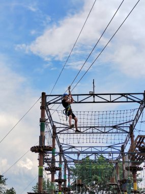 Spor kasklı ve güvenlik teçhizatlı genç bir çocuk halatla lunaparktaki teleferikle gökyüzünün arka planına doğru iniyor.