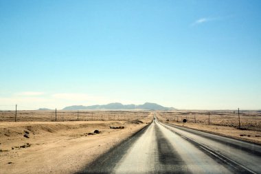 Çölde çok uzun bir yol var. Kenarlarında kum ve elektrik direkleri var. Uzakta küçük dağlar var. Sıcak iklim ve çöl manzarası