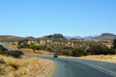 Bir araba, çöl bölgesinde perspektif olarak uzun ve dolambaçlı bir yol boyunca gider. Uzakta küçük dağlar var. Resimli bir turist rotası.