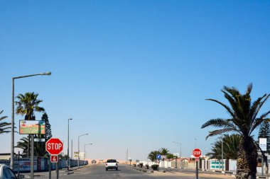 Küçük bir kasabadaki yola bakış açısı. İleride bir araba gidiyor ve kenarlarda yol işaretleri ve mavi gökyüzünün arka planında büyüyen bir palmiye ağacı var.