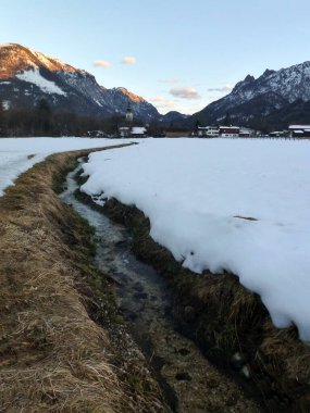 Dondurulmamış küçük bir nehir, bir kar tarlasından dağlara doğru akar mavi bir gökyüzüne karşı.