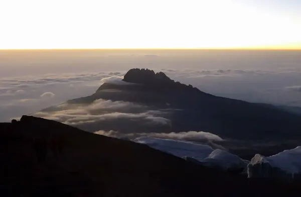 Top view of the mountain peak with clouds creeping over it against the background of the sunset and the distant horizon line