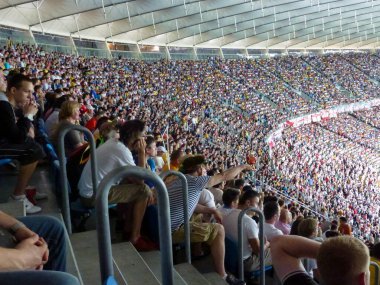 Büyük bir futbol stadyumunda seyirci ve taraftarlarla sıra sıra dizilmiş. Etkinlik ve spor