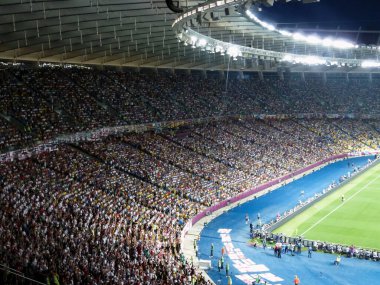 Büyük bir futbol stadyumunda seyirci ve taraftarlarla sıra sıra dizilmiş. Etkinlik ve spor