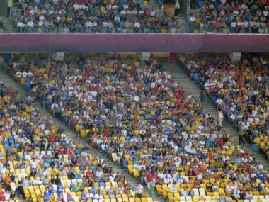 Büyük bir futbol stadyumunda seyirci ve taraftarlarla sıra sıra dizilmiş. Etkinlik ve spor