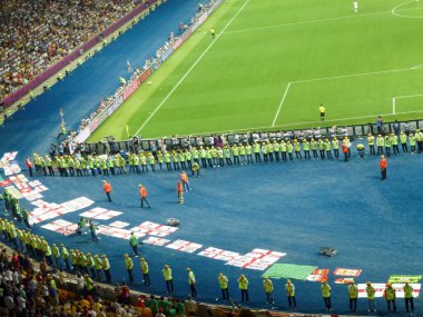 Büyük bir futbol stadyumunda seyirci ve taraftarlarla sıra sıra dizilmiş. Etkinlik ve spor