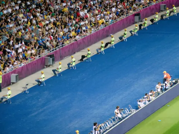 stock image At the football stadium, a number of volunteers sit on chairs in front of the stands with spectators and fans