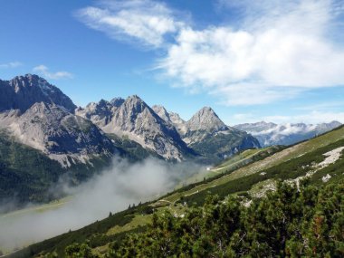 Mavi ufku olan yüksek bir dağ. Ön planda, yamaç yeşil orman ve bulutlarla kaplıdır. Güzel doğal manzara