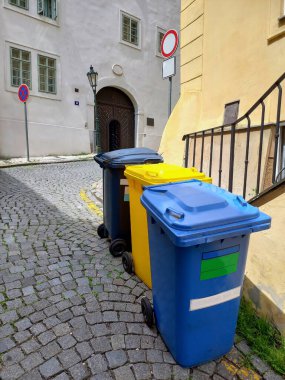 Farklı türde çöp toplamak için üç farklı renkte çöp kutusu. Taştan bir kaldırım taşının üzerindeki eski bir evin korkuluğunun arkasında duruyorum.