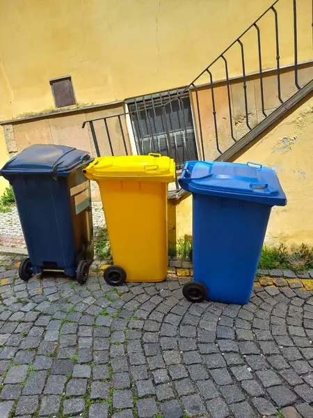 Three multi-colored garbage bins for collecting different types of garbage. The railings of an old house stand in the background