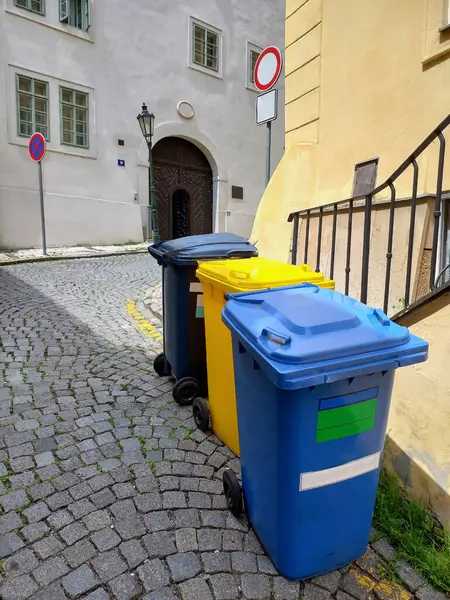 Three multi-colored garbage bins for collecting different types of garbage. Standing on the background of the railing of an old house on a stone cobblestone