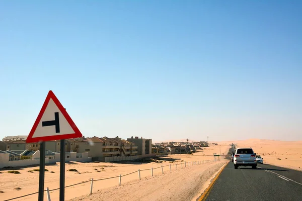 Cars drive along a desert road in perspective. On the side are the buildings of a small town against the background of the blue sky