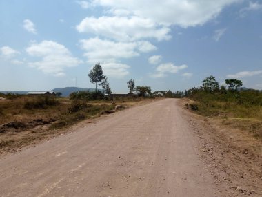 Çöl arazisindeki geniş toprak yol alçak ağaçlar, mavi gökyüzü ve köyün alçak binalarına karşı perspektiftir.