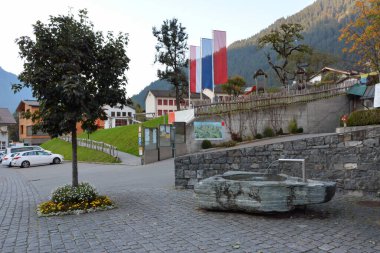 Otelin yakınına, dağın altına park etmiş. Ön planda dekoratif bir su kaynağı ve kaldırımda bir ağaç var.