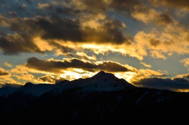 Karanlık bulutlarla kaplı akşam gökyüzü, parlak güneşin arka planında, dağların ardında batıyor. Işıklar bulutların arasından görülebilir.