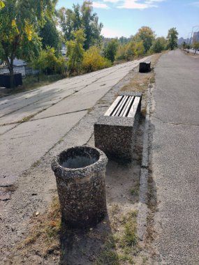 Taş süslemeli çöp kutusu ve kaldırım kenarındaki bankta mavi gökyüzüne ve ağaçlara bakış açısıyla
