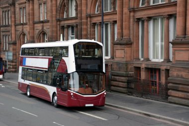 Arka planda tarihi bir bina bulunan çift katlı bir otobüs Londra, İngiltere 'de bir caddeden geçiyor. Turistler manzarayı inceliyor