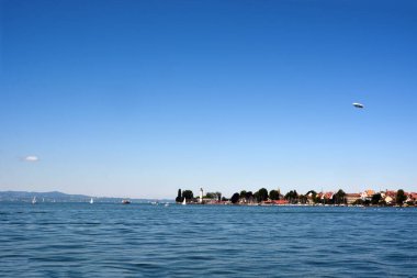 Uzak sahilde deniz manzaralı ve setin üzerinde antik evler var. Yatlar kıyıya yakın yüzer. Gökyüzünde bir hava gemisi uçuyor