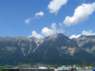 Küçük bir kasabanın üzerindeki orman dağlarının ve beyaz bulutlu mavi gökyüzünün resmi.