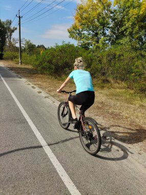 Genç bir kadın parktaki asfalt yolda bisiklet sürüyor. Doğada aktif bir eğlence. Ağaçların arka planında arka görünüm