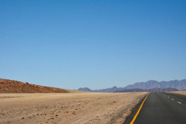 Açık mavi gökyüzünün altında çölde asfalt yol perspektifi. Yol uzak dağlara ve tepelere çıkar.