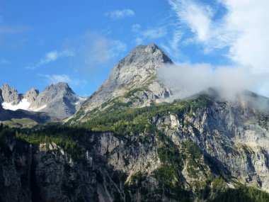 Yeşil ormanlarla kaplı kayalık dağ yamaçlarının manzarası. Üzerlerinde mavi bir gökyüzü ve beyaz bulutlar var.