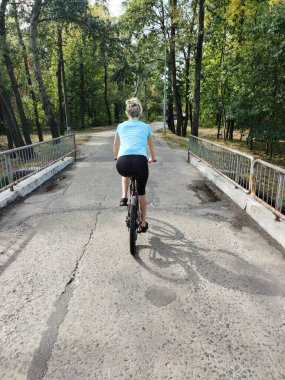 Genç bir kadın, parktaki bir ara sokakta, korkuluklu bir köprünün üzerinde bisiklet sürüyor. Bakış açısına göre. Aktif ve spor yaşam tarzı