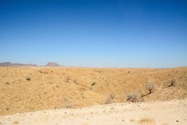 Kuru çöl manzarası, ufukta küçük bitkiler ve alçak tepeler, mavi gökyüzünün altında.