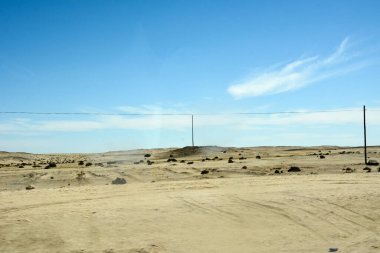 Kuru bir çölde sıcak bir manzara. Elektrik direkleri ön planda. Arka planda uzak dağlar ve açık mavi bir gökyüzü var.