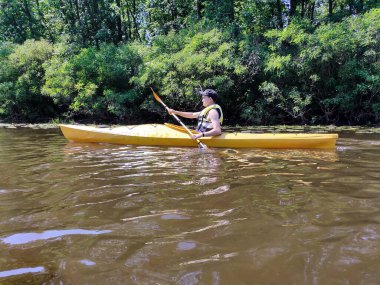 Genç bir Kafkasyalı adam, orman ağaçlarının arka planına karşı nehirde bir kanoyla yüzüyor. Yan görüş. Spor ve aktif bir yaşam tarzı