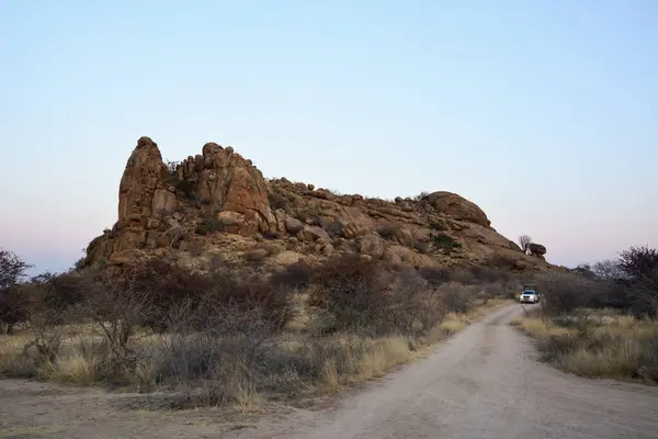 stock image Near a large rocky hill made of large boulders, a dirt road is laid along which a car drives against the background of a blue sky. Desert landscape and wild nature