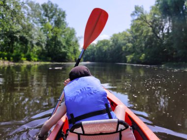 Panama giymiş bir çocuk dar bir nehirde kanoyla yüzer ve kürek çeker. Dikiz aynası. Yaz sporları ve eğlence