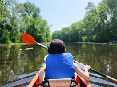 Panama giymiş bir çocuk dar bir nehirde kanoyla yüzer ve kürek çeker. Dikiz aynası. Yaz sporları ve eğlence