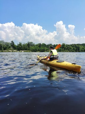 Şapkalı genç bir beyaz adam sahildeki yeşil bir ormanın arka planında kanoyla yüzüyor. Yaz sporları ve eğlence