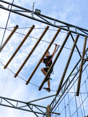 Asma halat köprüsünde ilerleyen bir gencin tırmanma eğitimi. Aşağıdan, gökyüzünün arka planından görüntüle. Çocuklar ve gençlerin spor eğitimi