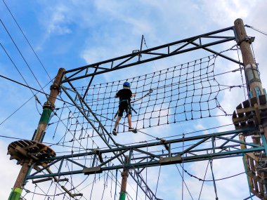 Asma halat köprüsünde ilerleyen bir gencin tırmanma eğitimi. Aşağıdan, gökyüzünün arka planından görüntüle. Çocuklar ve gençlerin spor eğitimi