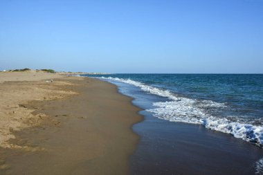 Işık dalgaları geniş bir deniz sahiline doğru akmaktadır. Mavi berrak gökyüzü altında doğal yaz turistik manzarası