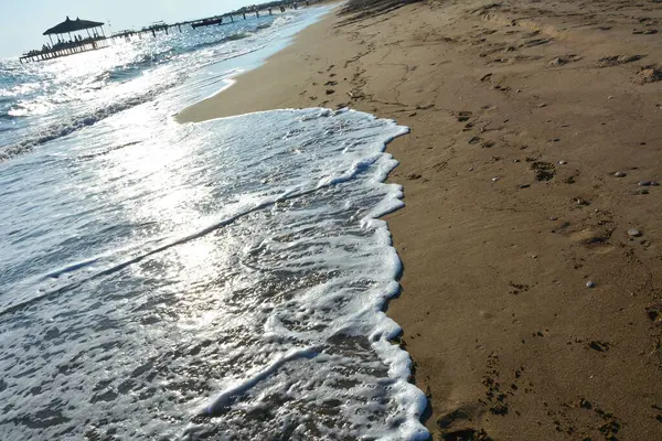 Işık dalgaları geniş bir deniz sahiline doğru akmaktadır. Mavi berrak gökyüzü altında doğal yaz turistik manzarası